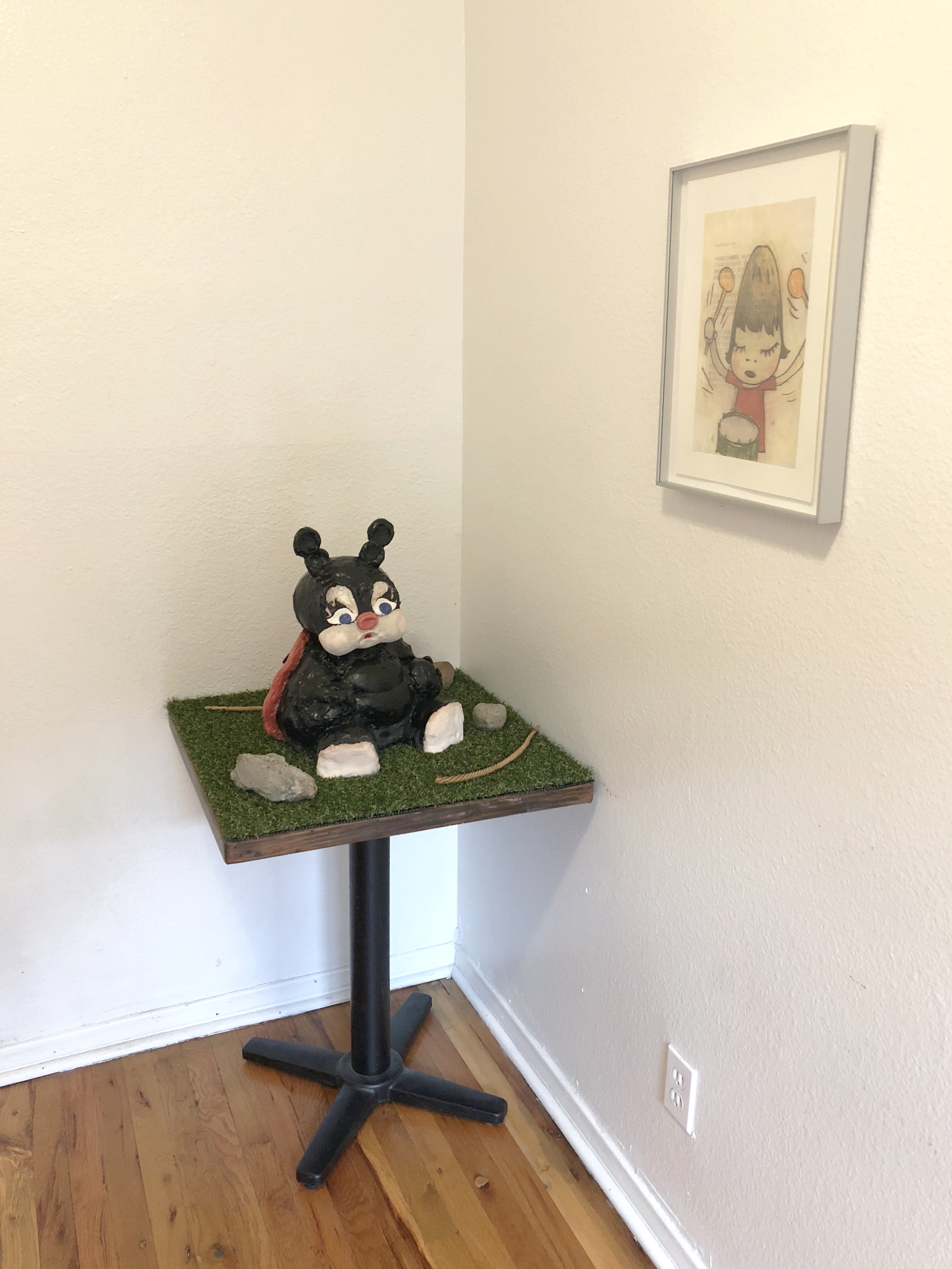 A ceramic sculpture of a black ladybugy on a small table covered in astroturf with a drawing hung on the wall beside it